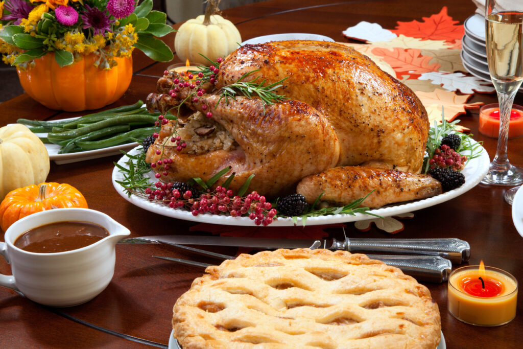 Roasted pepper turkey for Thanksgiving, garnished with pink pepper, blackberry, and fresh rosemary twigs on a dinner table decorated with mini pumpkins, beans, carrots, baked potato, pie, cranberry relish, gravy, flowers, candles, and flutes of champagne, Holiday Braces & Aligners Care: Keep Your Smile Safe During Festive Feasts