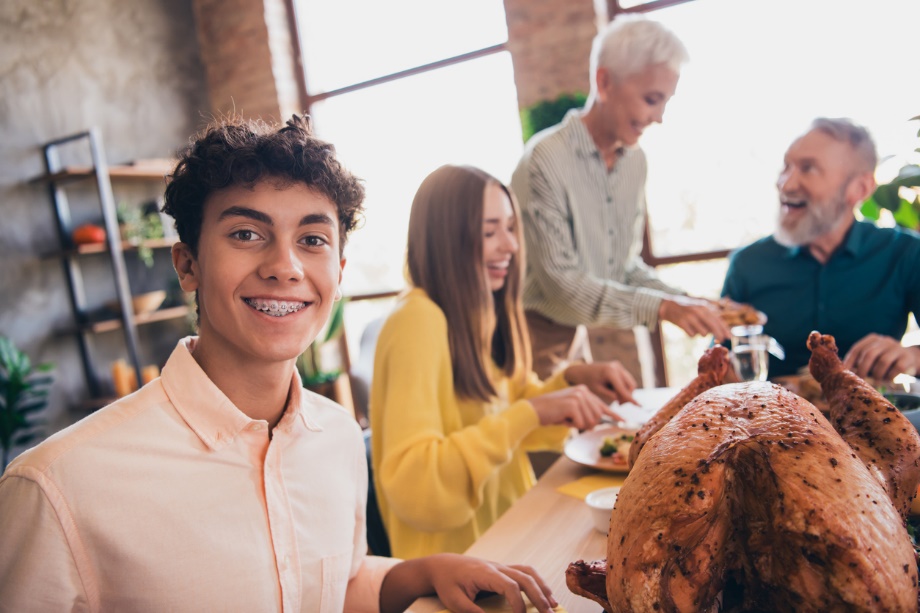 a young person smiling in the foregound, A group of people eating food in the background, Holiday Braces & Aligners Care