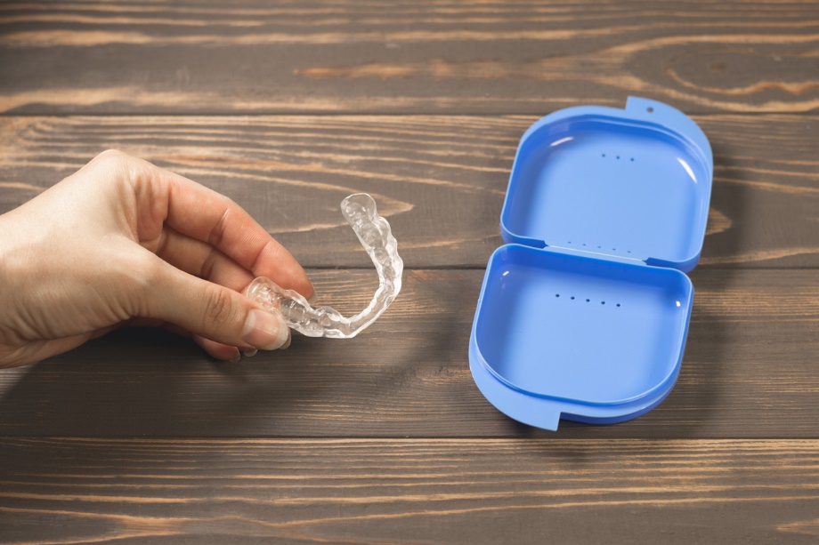 A hand holding a clear plastic retainer, over a plastic container, Holiday Braces & Aligners Care