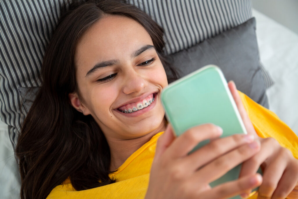 Happy teen girl with braces using her smartphone lying in bed smiling